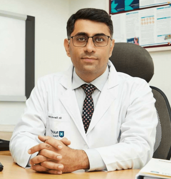 Male doctor wearing a white coat and glasses sits at a desk in a clinic, with a computer showing medical graphics.