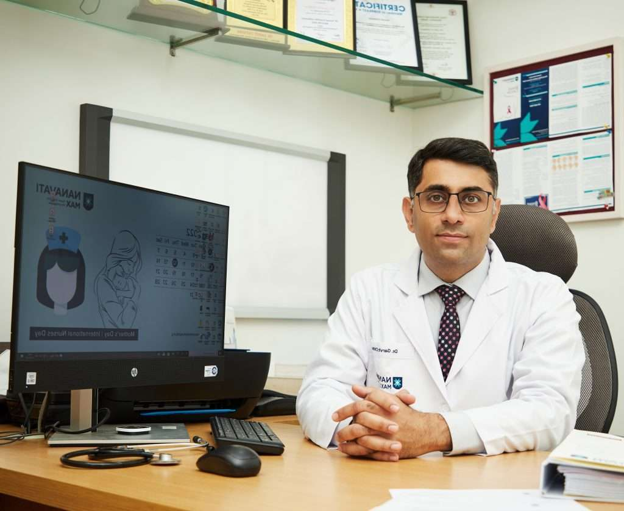 Picture3 | Dr Garvit Male doctor in a white coat sits at a desk in a clinic, hands folded, with a computer monitor displaying a medical illustration behind him and framed certificates on the wall.