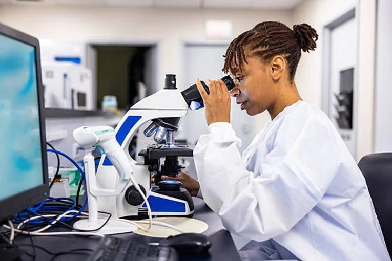 Young female researcher in a white lab coat looks through a microscope at a lab bench.